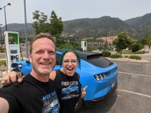 Smiling man and woman in front of blue Mach-E electric vehicle connected to an EV charging station.